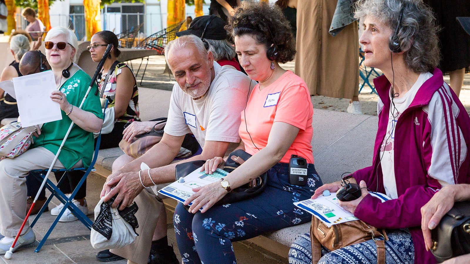 People in colorful outfits sit on a bench holding tactile maps and wearing headphones listening to a verbal description tour.